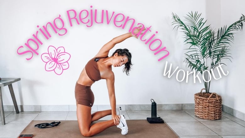 A woman wearing a sports bra and bike shorts, kneeling and stretching backward on an exercise mat in a living room. A planted palm tree is behind her. Words "spring rejuvenation workout" are on screen.