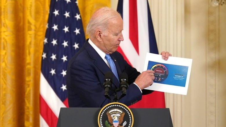 Joe Biden, the president of the united states, who is wearing a dark suit and light blue tie, stands behind a lectern with the presidential seal and examines a chart printed on a piece of paper he holds in his hands. 