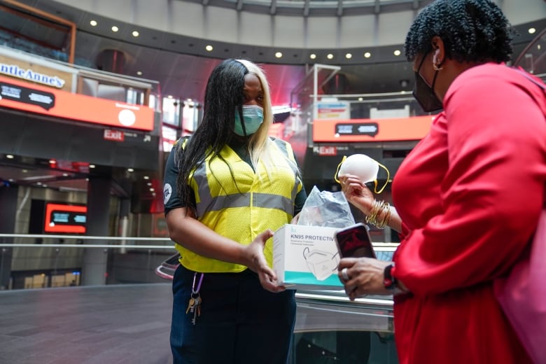 A woman in a mask and a reflective vest hands out a mask to a person.