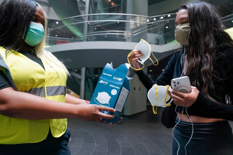 A woman wearing a neon yellow reflective vest and a mask holds out a box of N95 masks as another masked woman picks out two masks and holds a phone. 