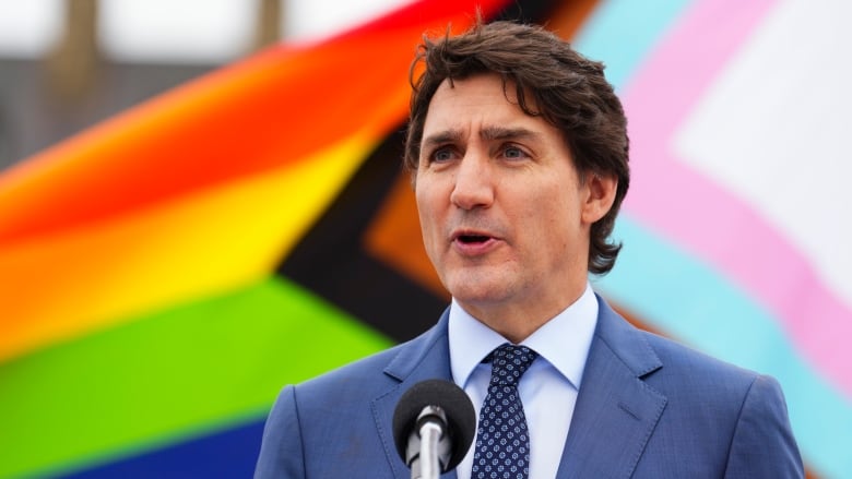 A man in a suit stands in front of a rainbow flag.