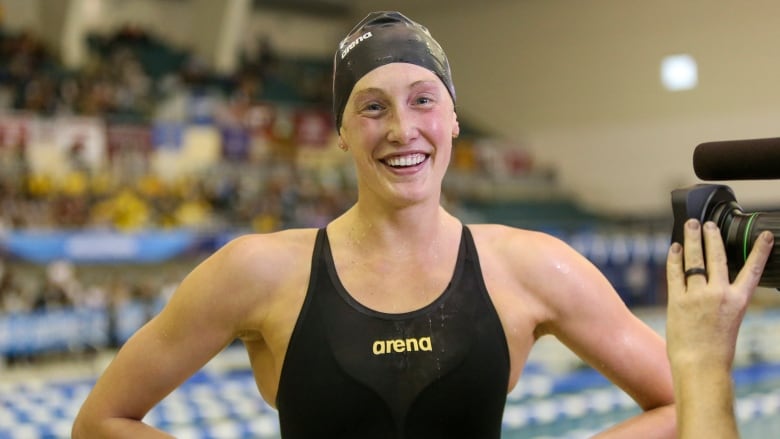 A swimmer in a black swim suit and smin cap smiles as she stands outside of a pool.